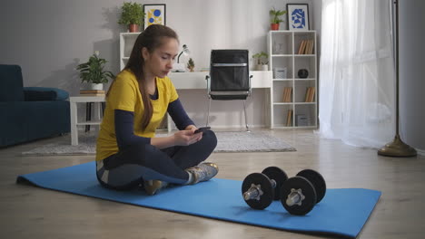 young-woman-is-resting-on-sporty-mat-with-dumbbells-using-smartphone-for-surfing-internet-workout-at-home-social-net-addiction